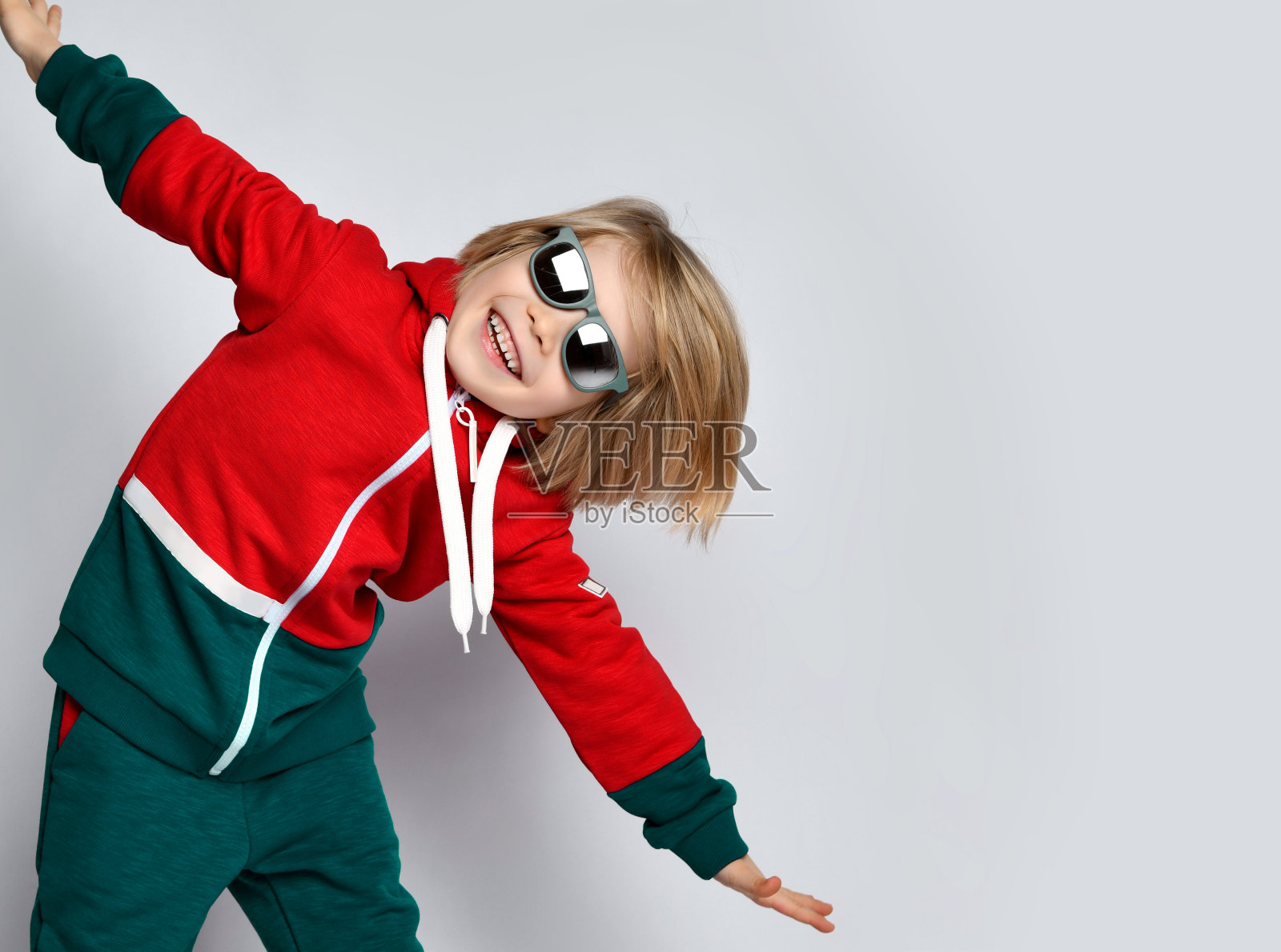 微笑积极活泼的金发男孩穿着深灰色和红色的连帽衫，裤子和太阳镜扮演飞机飞行员照片摄影图片