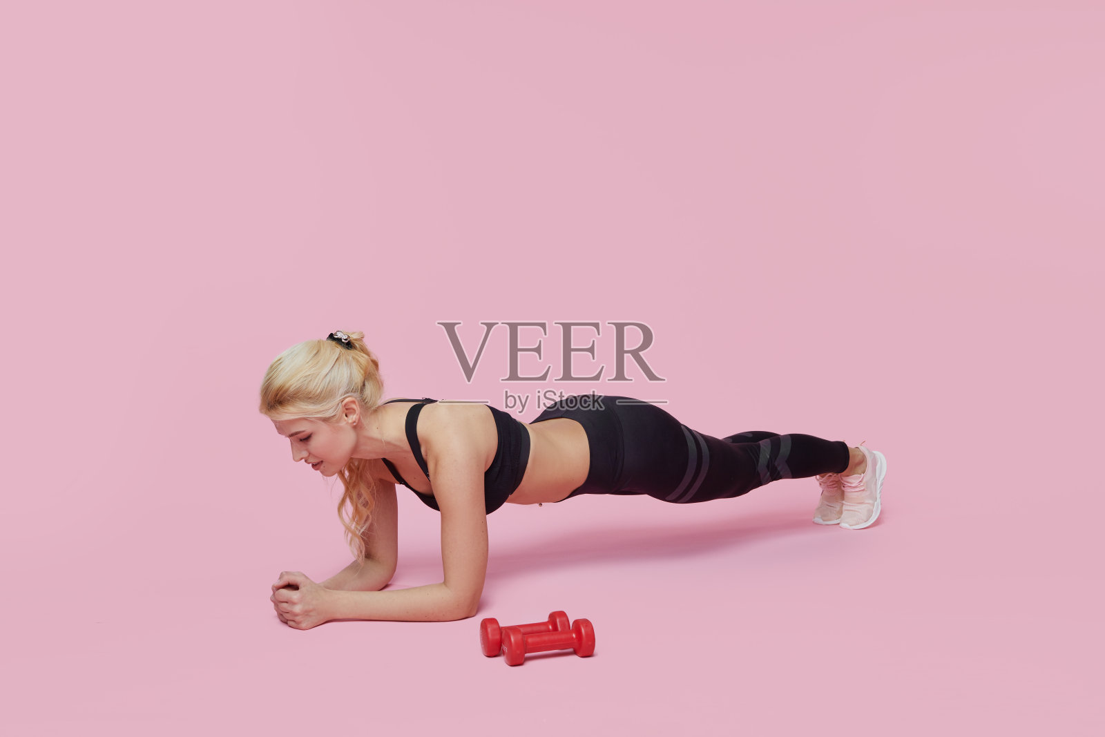 身材苗条的健身女孩在粉色背景上用哑铃训练肌肉照片摄影图片
