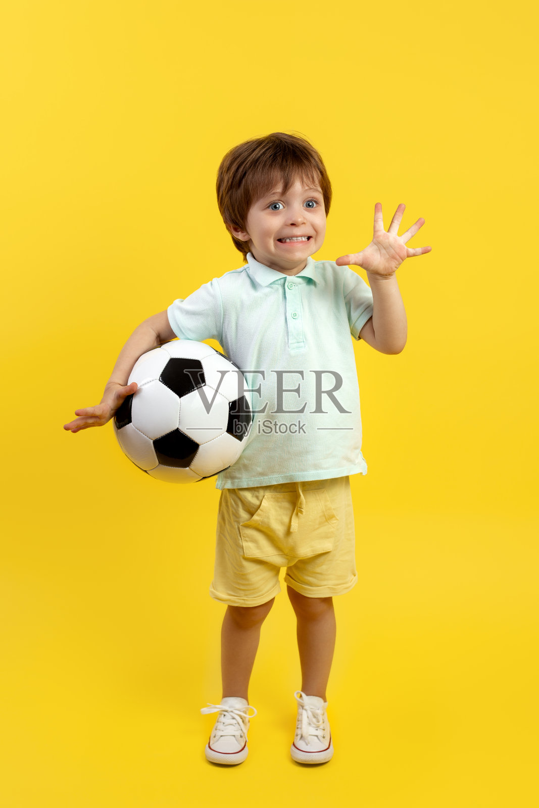可爱的小男孩穿着休闲的夏装，拿着一个足球，在黄色的背景上摆出滑稽的脸。照片摄影图片