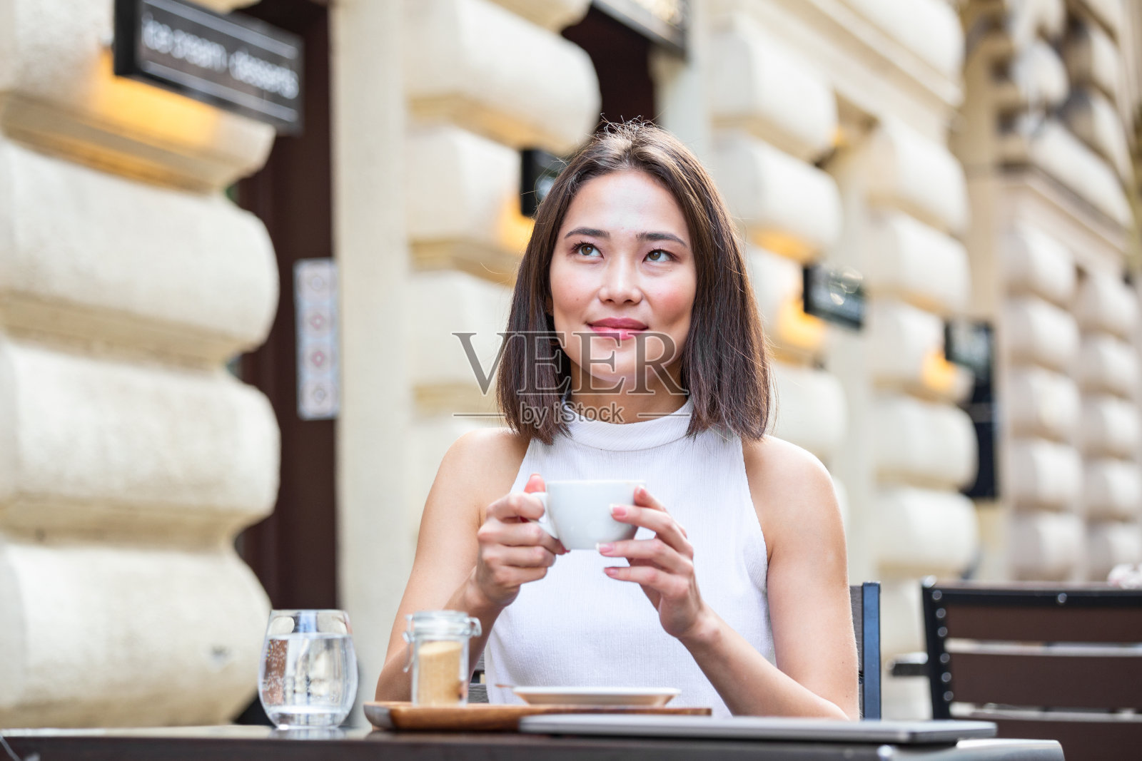 微笑的亚洲女子喝着咖啡，使用手机。满意的女子享受着一杯咖啡。美丽的女孩在咖啡店里用白色马克杯喝咖啡的特写照片摄影图片