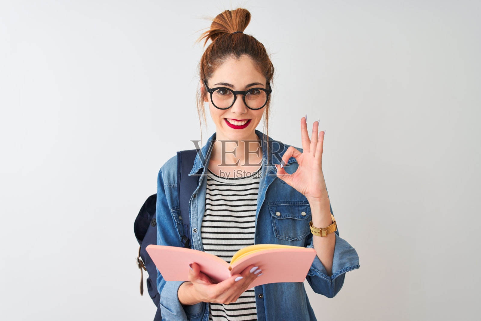 红发女学生背着书包，在白色背景上读书，用手指做OK手势，象征优秀照片摄影图片