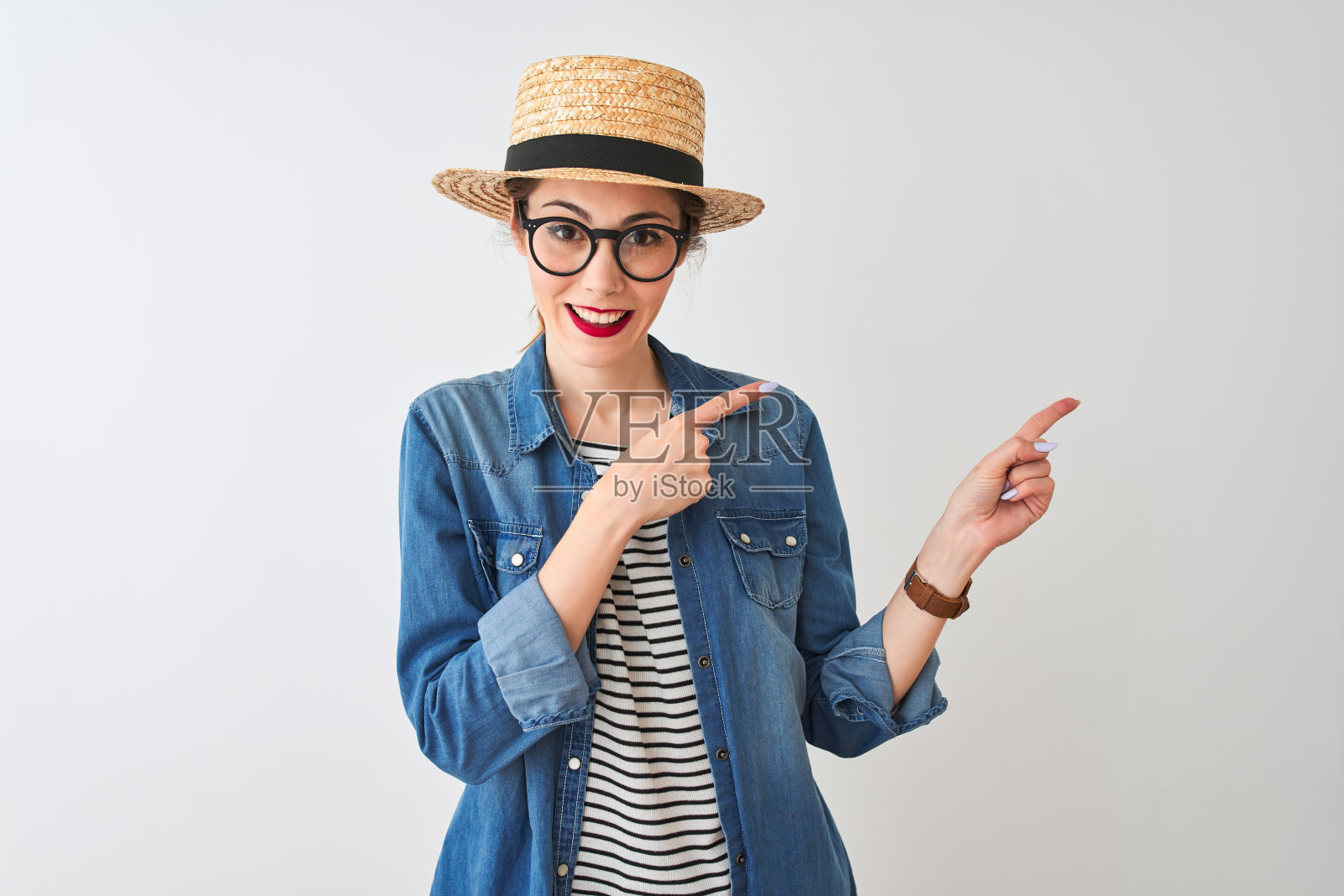 红发女人戴着牛仔衬衫、眼镜和帽子，在白色背景前微笑地看着镜头，用双手和手指指向侧面照片摄影图片