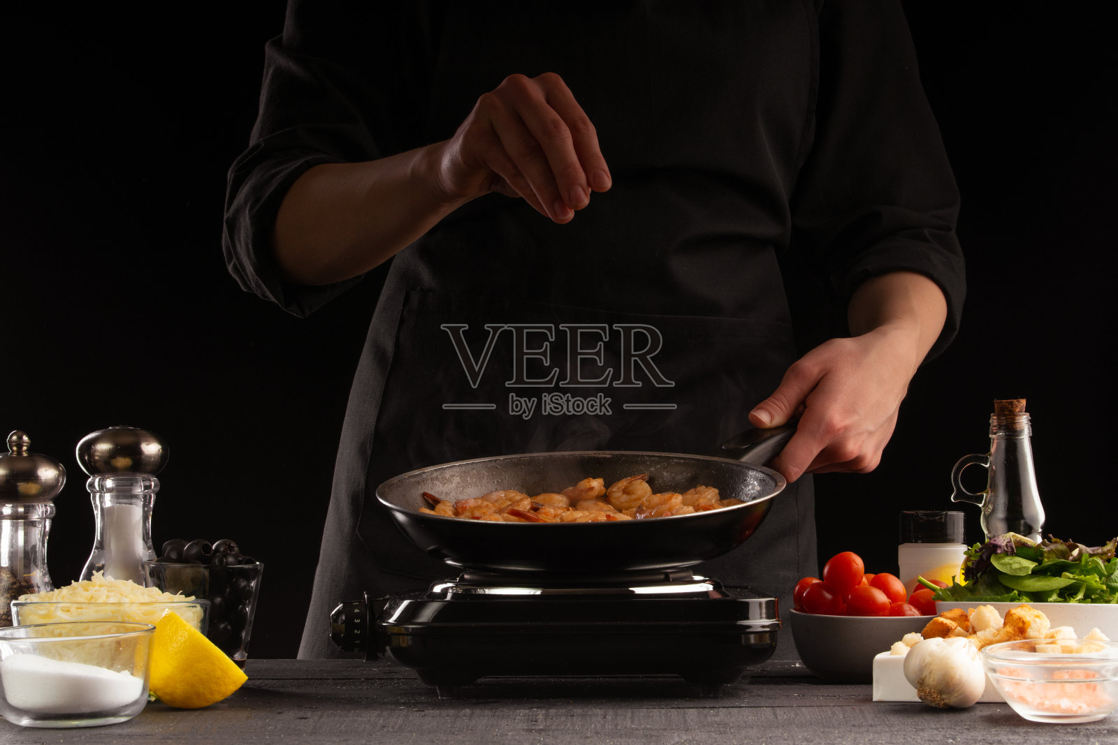 厨师将海鲜、虾，撒上盐，在煎锅中炸熟，动态冷冻。以蔬菜为背景。黑色背景，烹饪和食谱，烹饪和健康饮食。照片摄影图片