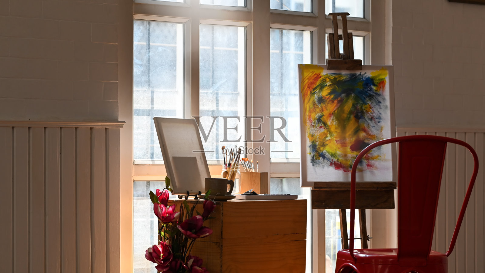 晨光中艺术工作室的照片。椅子，木桌，油画布与画架，画框，花，花瓶，咖啡杯，笔筒，画笔，窗户。凌乱的艺术家工作场所。照片摄影图片