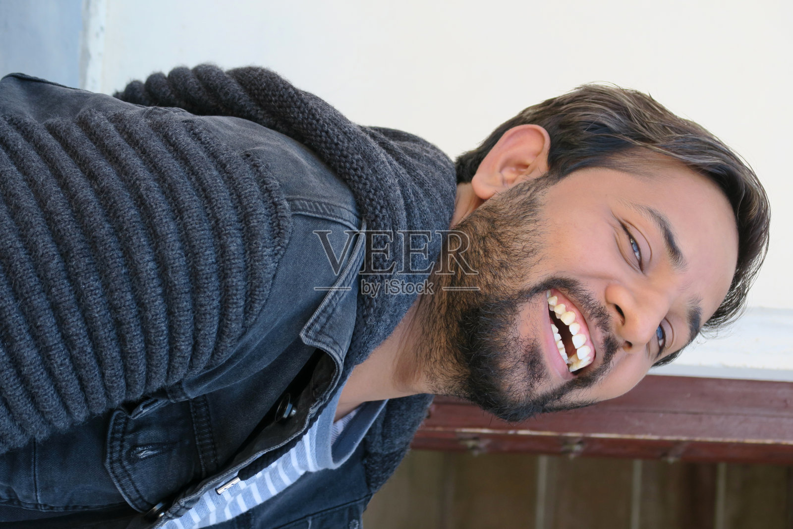 年轻英俊的印度男子傻乎乎地向后仰着头大笑，快乐的印度男子在20岁出头微笑着，假日坐在楼梯上开心地做鬼脸，男子的胡子乱弄，黑色的帽衫，蓝色的白色t恤。照片摄影图片