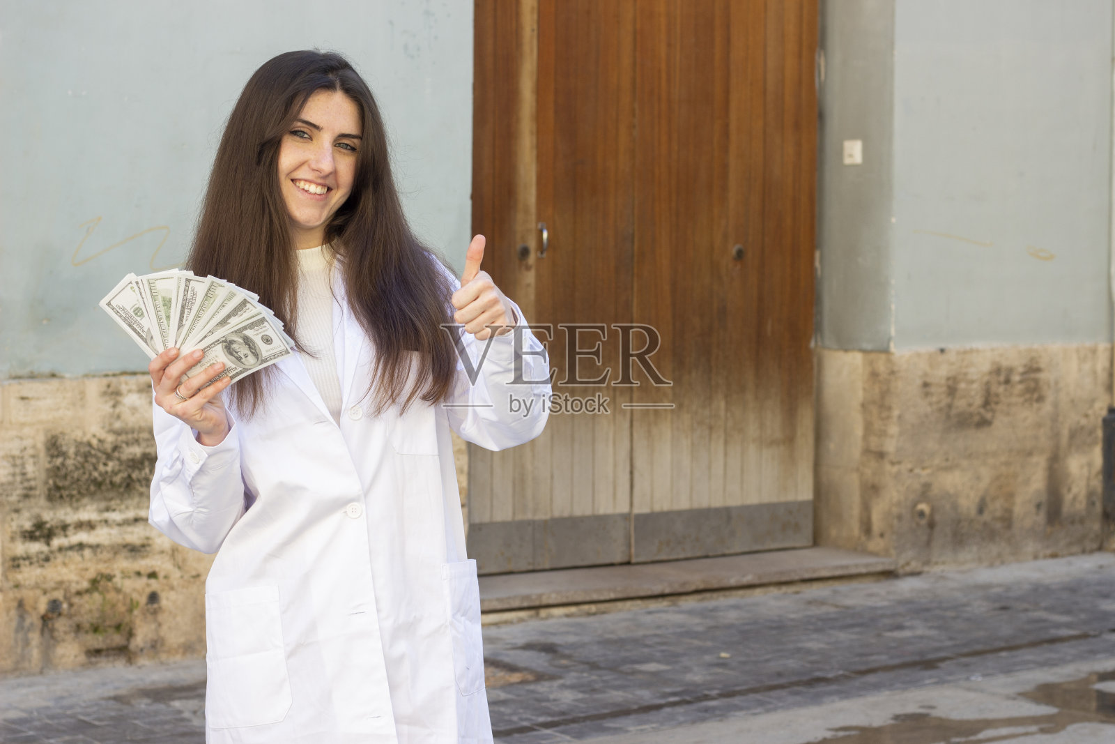 有吸引力的白人年轻女子的肖像模特显示拇指向上的手势与美元钞票。护士，医生，科学家，治疗师，药剂师，在城市里，穿着白色大褂制服，黑暗的空气。的地方照片摄影图片