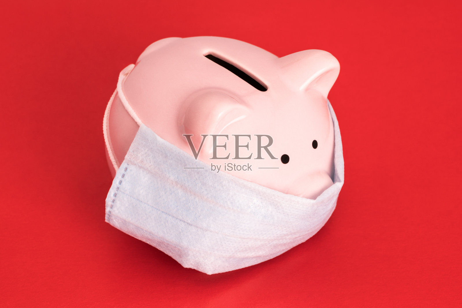 粉红色的小猪储蓄罐站在大红色的背景上，戴着医用口罩。COVID-19经济危机照片摄影图片