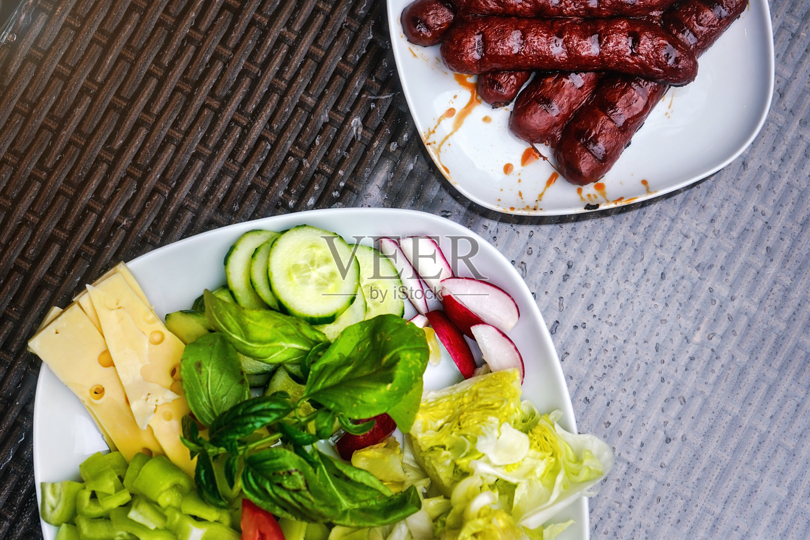 两个白色盘子，一个放烤香肠/法兰克福香肠，另一个放沙拉——黄瓜，萝卜，罗勒，胡椒和奶酪。从上面的观点照片摄影图片