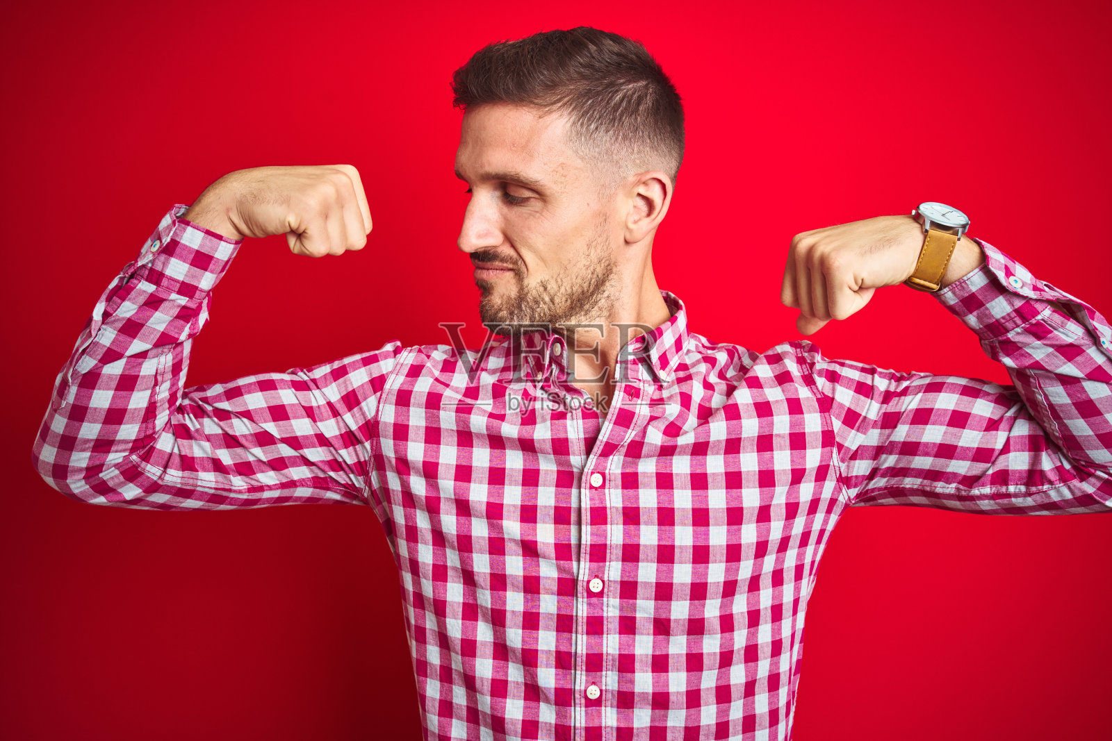 在红色背景下展示手臂肌肉，面带自豪微笑的年轻英俊男子，健身理念照片摄影图片