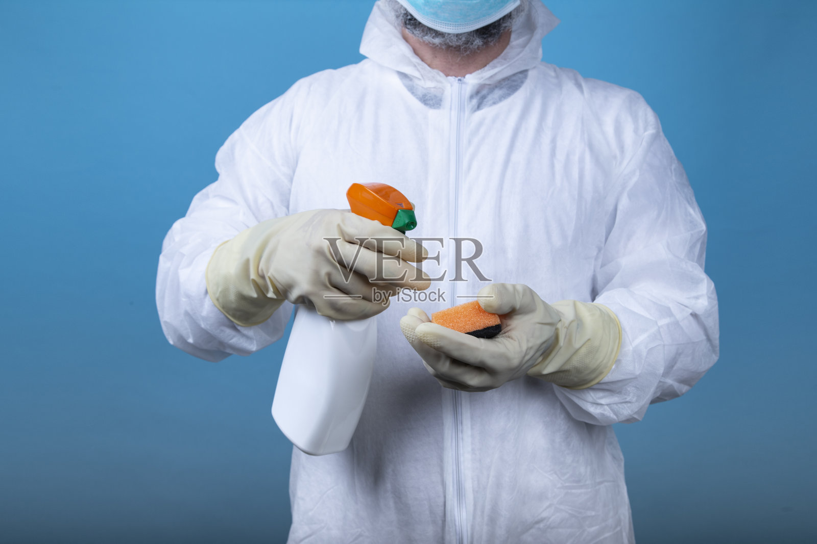 身着防护服和面罩的男子用白色的消毒液体喷雾和海绵在蓝色背景上。COVID-19冠状病毒的概念照片摄影图片