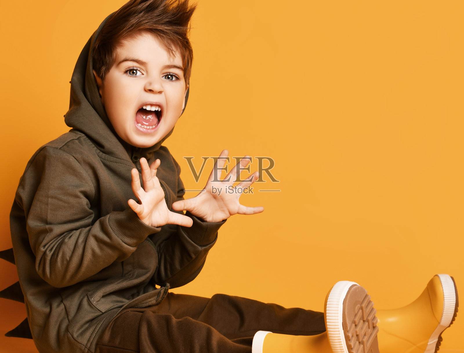 穿着棕色恐龙帽衫和裤子，黄色胶靴的男孩。坐在地板上，咆哮着吓唬你，以橙色为背景摆姿势照片摄影图片