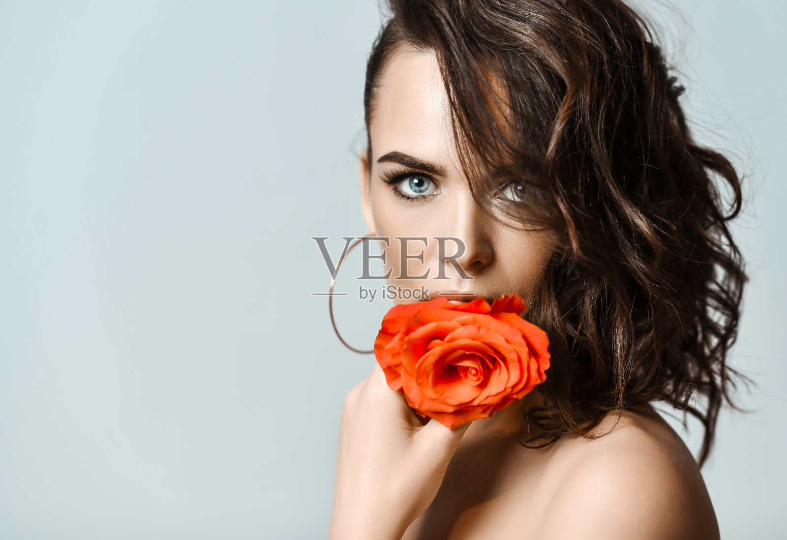 戴圆形耳环、手持玫瑰的性感年轻棕发美女肖像，浅灰色背景照片摄影图片