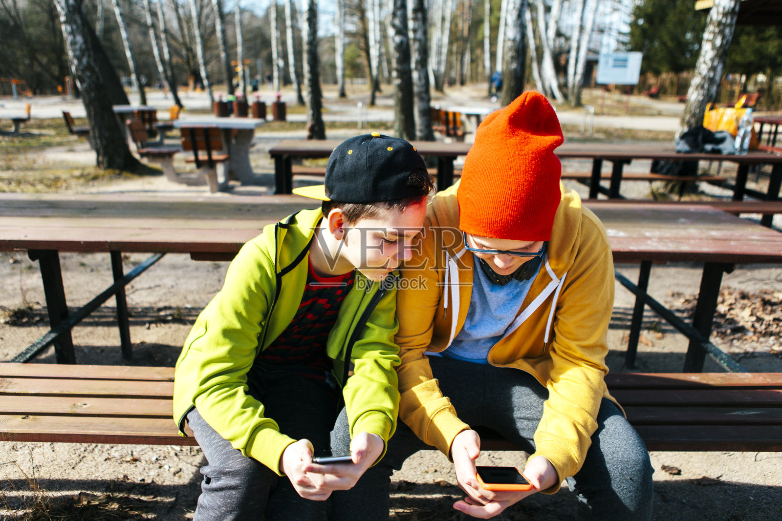 一个穿着黄色和绿色连帽衫坐在公园里阳光明媚的长椅上的男孩，兴致勃勃地从肩膀上看向弟弟的手机。照片摄影图片