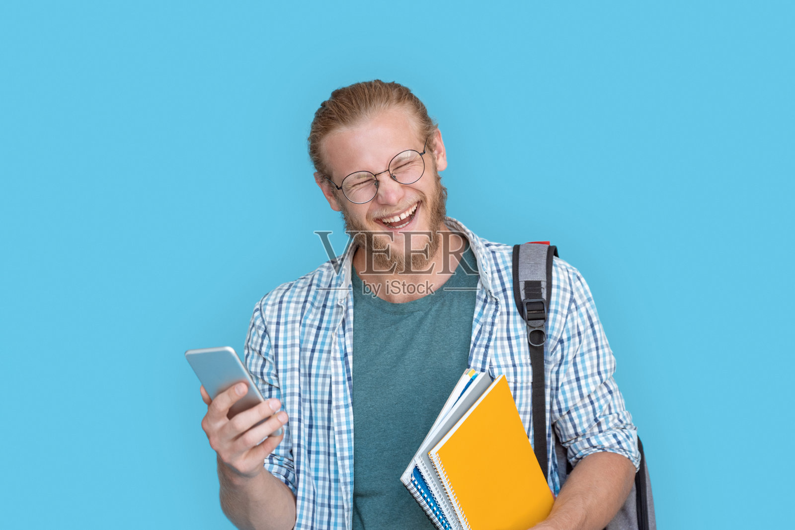 戴眼镜的快乐年轻男学生博主拿着智能手机，阅读短信，在聊天中嘲笑笑话，玩得开心，拍摄抖音视频博客，在线观看视频，蓝色工作室背景照片摄影图片