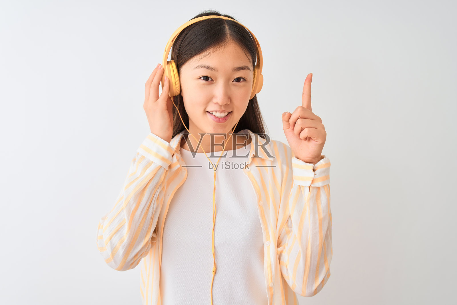 一名年轻的中国女子戴着耳机在孤立的白色背景下听音乐，惊讶地想到一个想法或问题，开心地指着对方的脸照片摄影图片