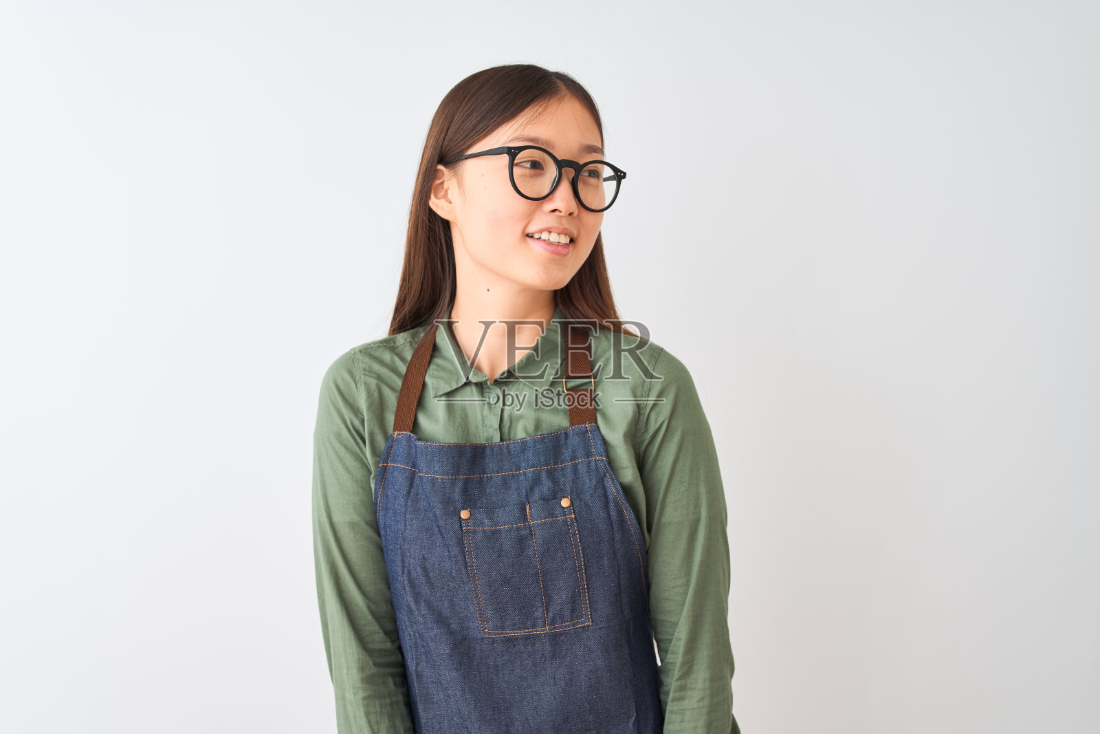 年轻的中国女店主穿着围裙和眼镜在孤立的白色背景看向一边，脸上带着微笑，自然的表情。自信的笑。照片摄影图片