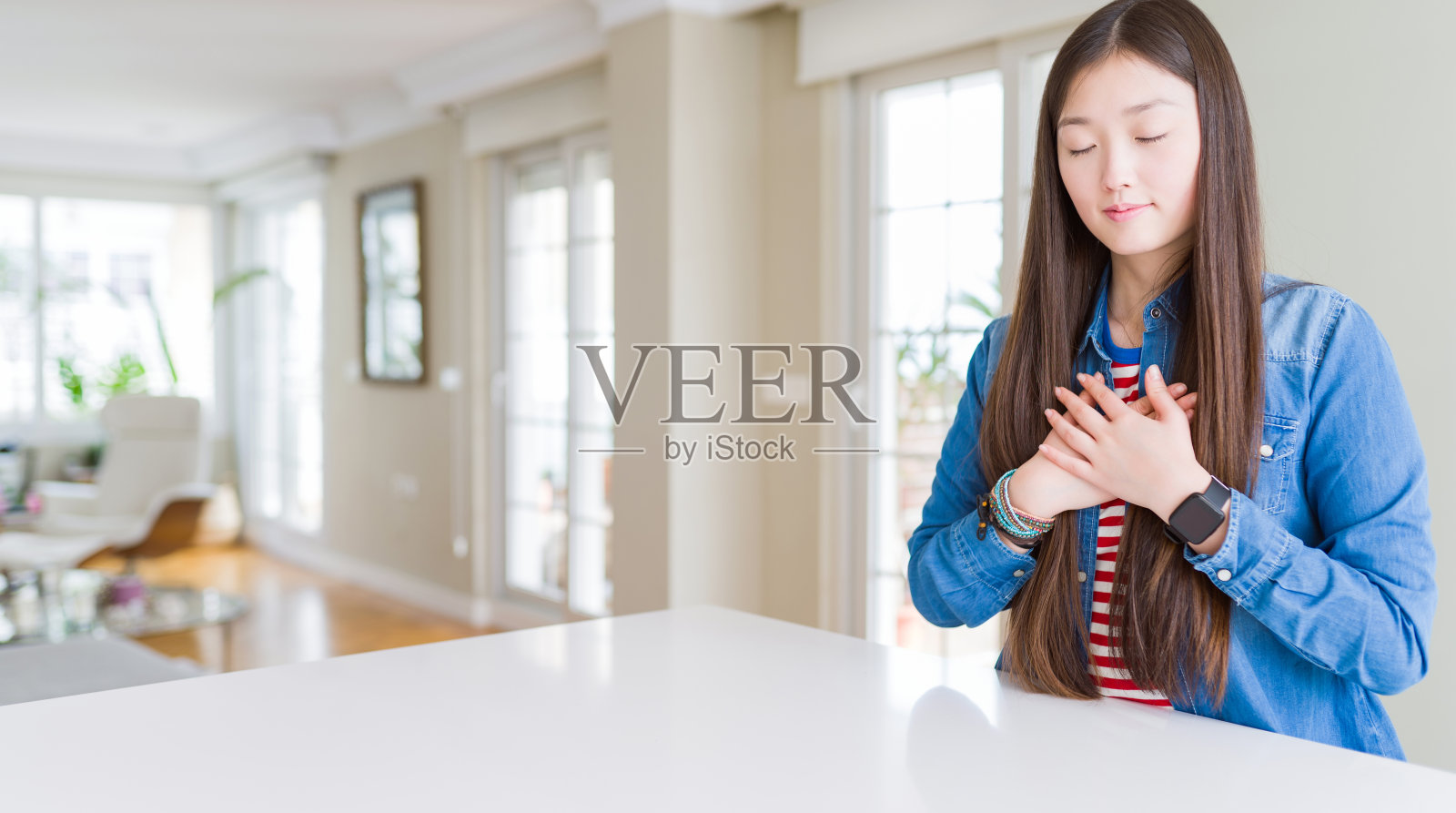 年轻美丽的亚洲女人与长头发穿着牛仔夹克微笑与双手在胸前闭上眼睛和感激的手势在脸上。健康的概念。照片摄影图片