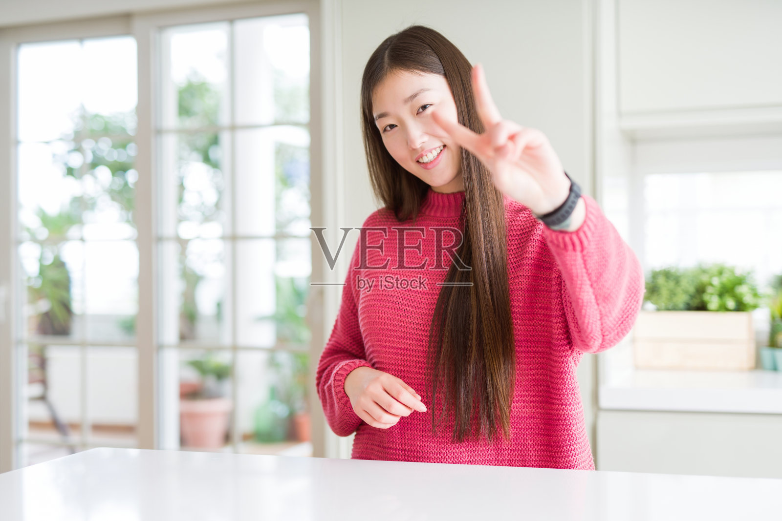 美丽的亚洲女人穿着粉红色的毛衣在白色的桌子上微笑看着镜头显示手指做胜利的手势。二号人物。照片摄影图片