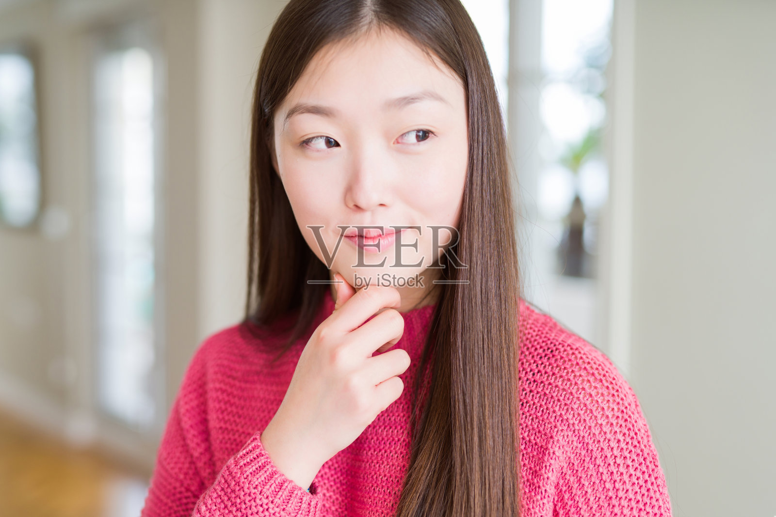 美丽的亚洲女人穿着粉红色的毛衣，手在下巴思考问题，沉思的表情。带着沉思的微笑。怀疑的概念。照片摄影图片