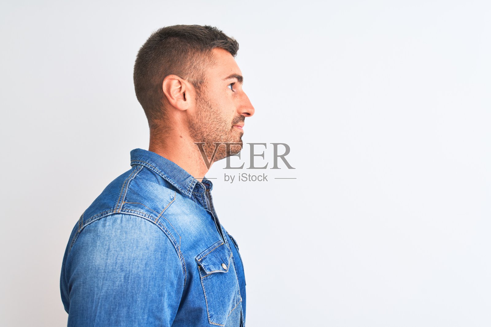 年轻英俊的男子穿着牛仔夹克站在孤立的背景看向一边，放松的侧面姿势与自然的脸与自信的微笑。照片摄影图片