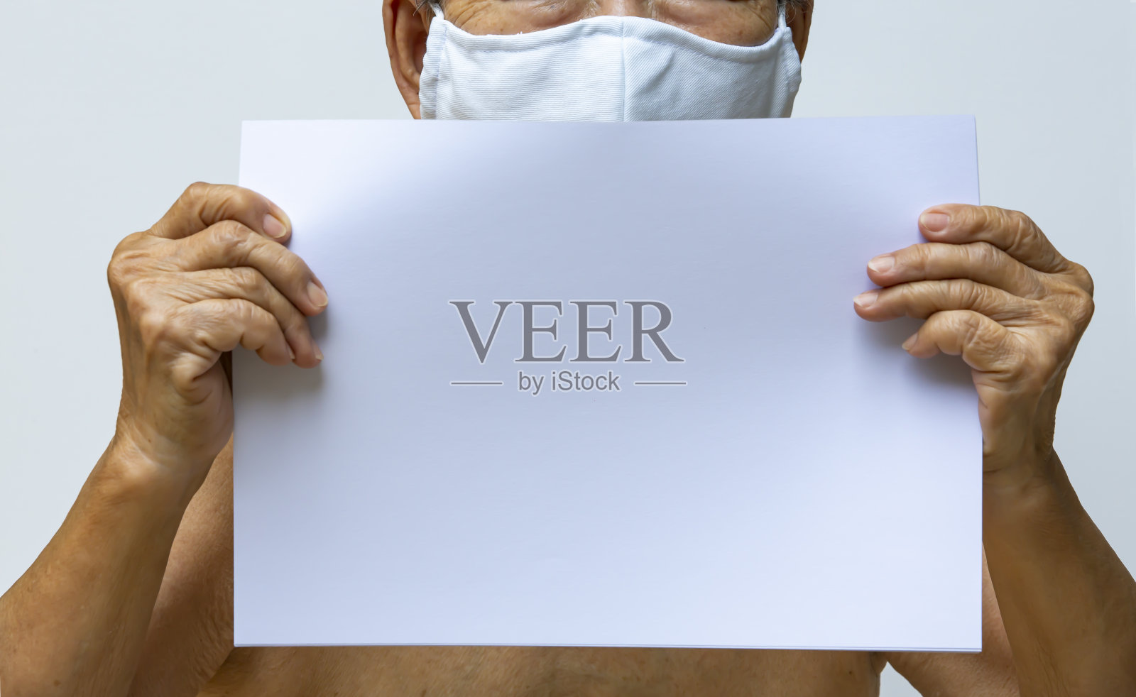 老年妇女手持空白纸a4，佩戴DIY手工布口罩、医用口罩、白色背景外科口罩、预防新冠肺炎、冠状病毒、细菌、医疗保健照片摄影图片