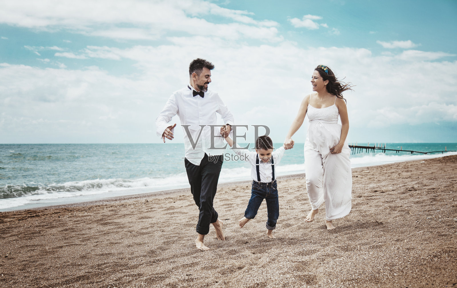 幸福的三口之家。在阳光明媚的日子里，怀孕的妈妈、爸爸和儿子在海滩上散步。照片摄影图片