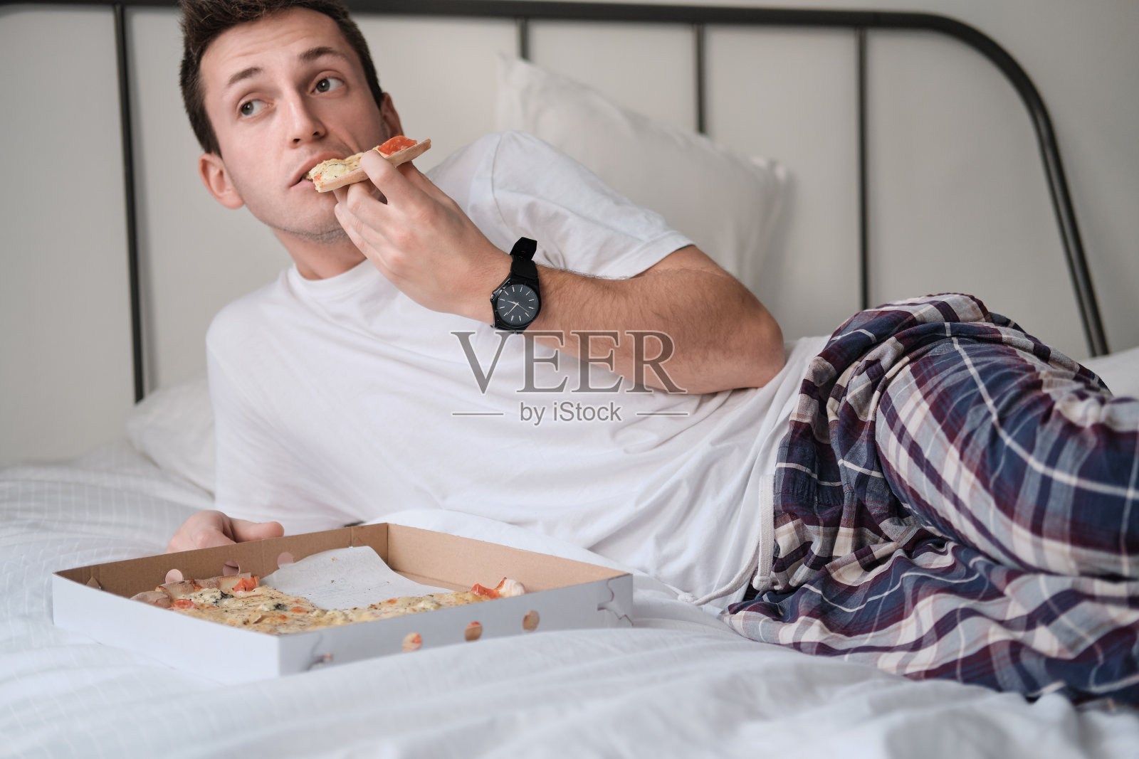 穿着白衬衫和睡衣的年轻人微笑着躺在床上，吃着美味的披萨。照片摄影图片