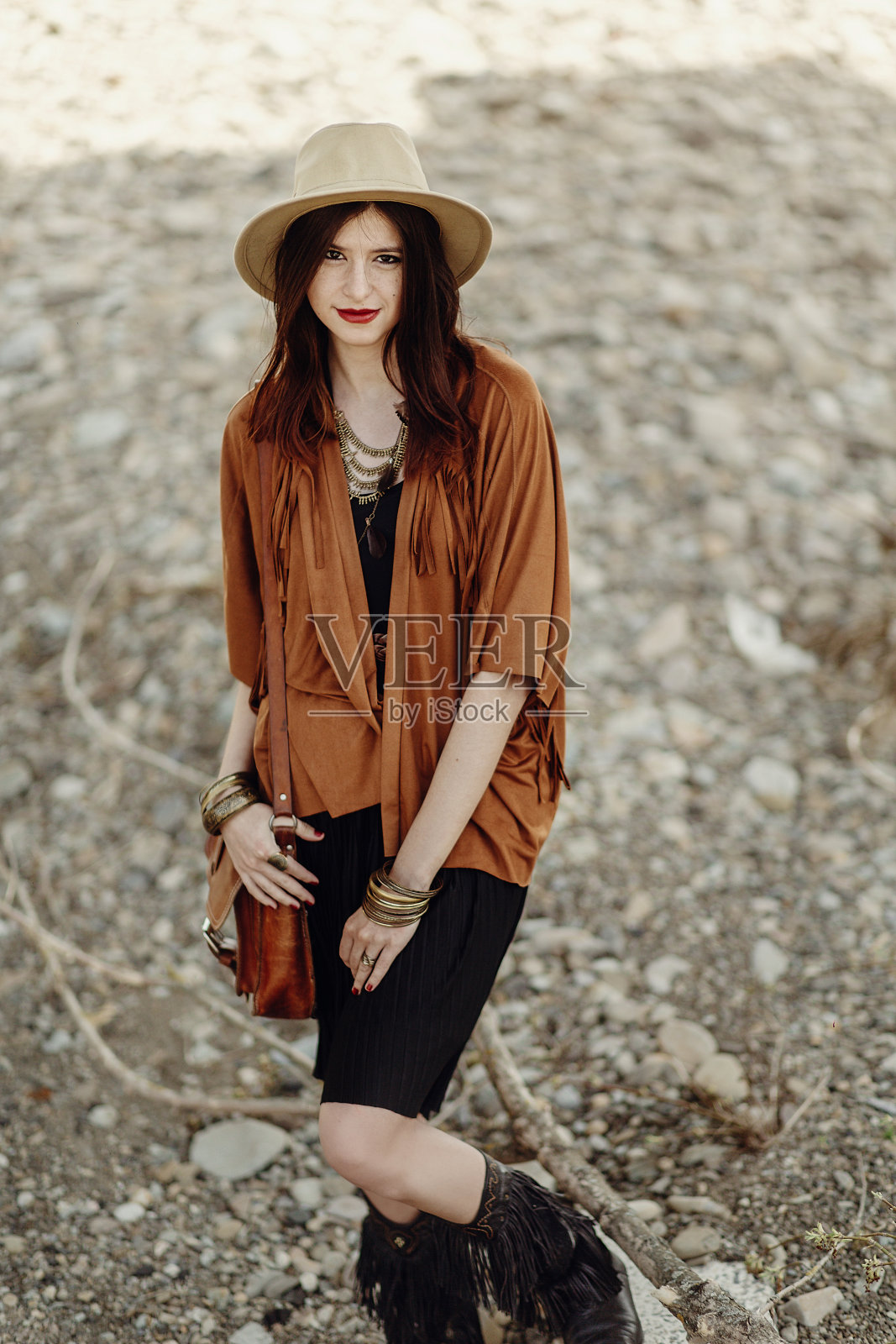 戴帽子、皮包、流苏披肩和靴子的时尚波西米亚风女人。在山区河流岩石附近摆姿势的年轻吉普赛嬉皮旅行者，迷人造型，氛围感照片摄影图片