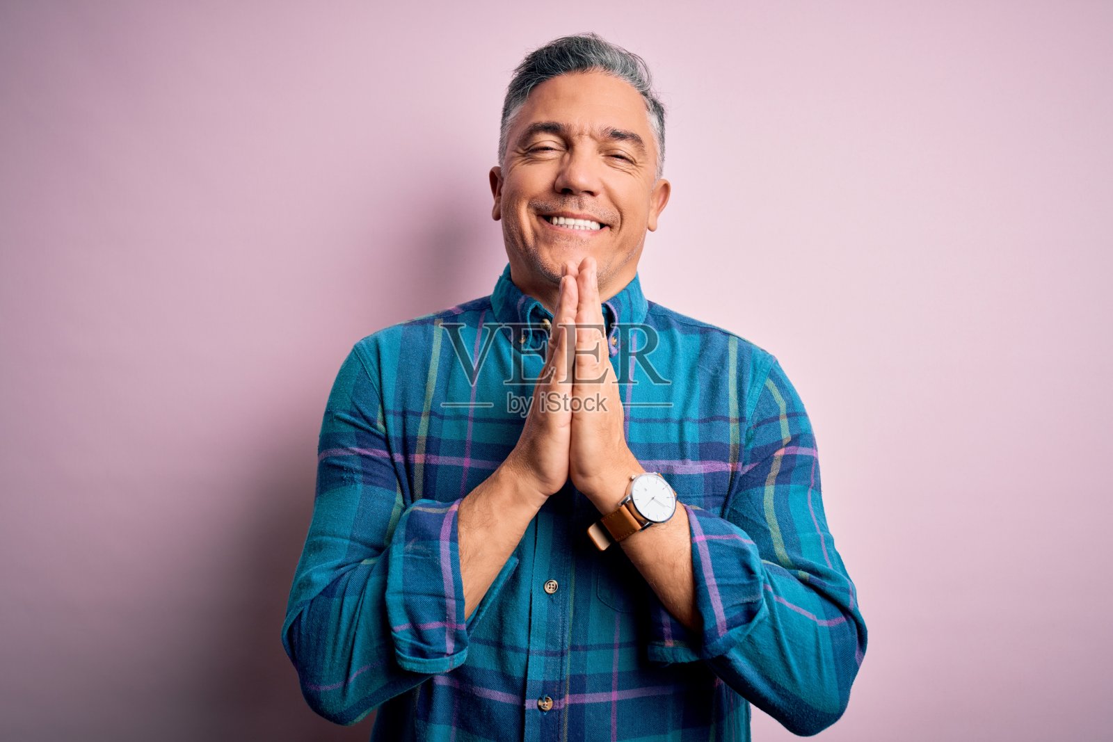 中年英俊灰发男子穿着休闲衬衫在粉红色背景前祈祷，双手合十请求原谅，面带自信的微笑照片摄影图片