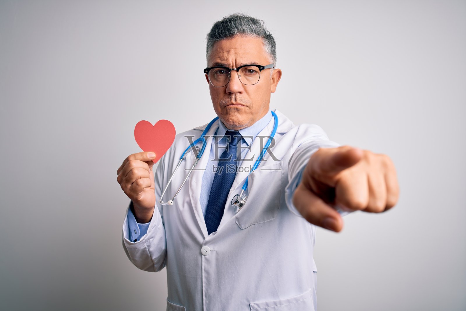 拿着红纸心的英俊灰发中年男医生指向镜头，传递积极自信的手势照片摄影图片