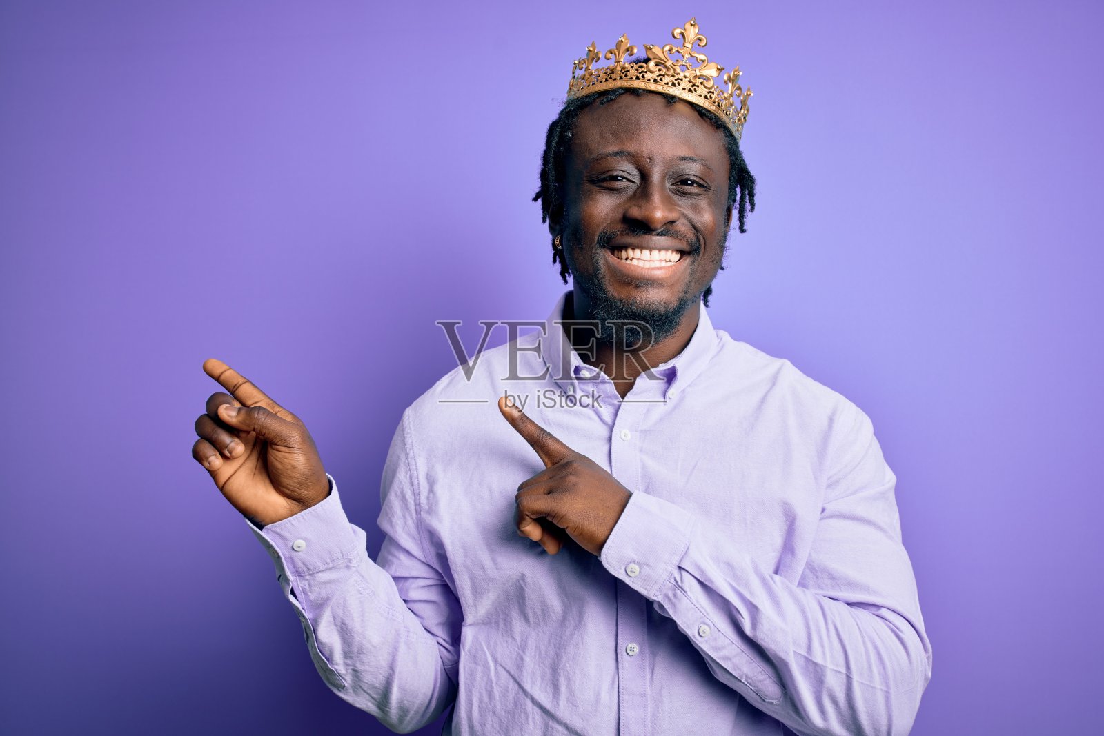 年轻的非洲裔美国人戴着金色的王冠，在孤立的紫色背景上微笑着看着镜头，双手和手指指向一边。照片摄影图片