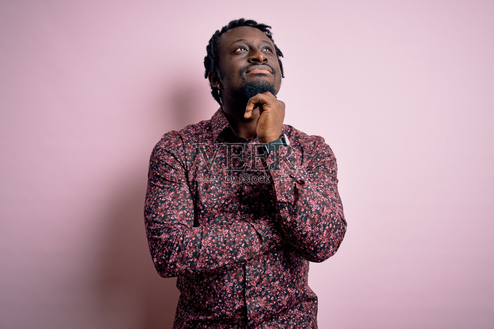 年轻英俊的非裔美国人穿着休闲衬衫站在粉红色的背景与手下巴思考问题，沉思的表情。微笑和沉思的脸。怀疑的概念。照片摄影图片
