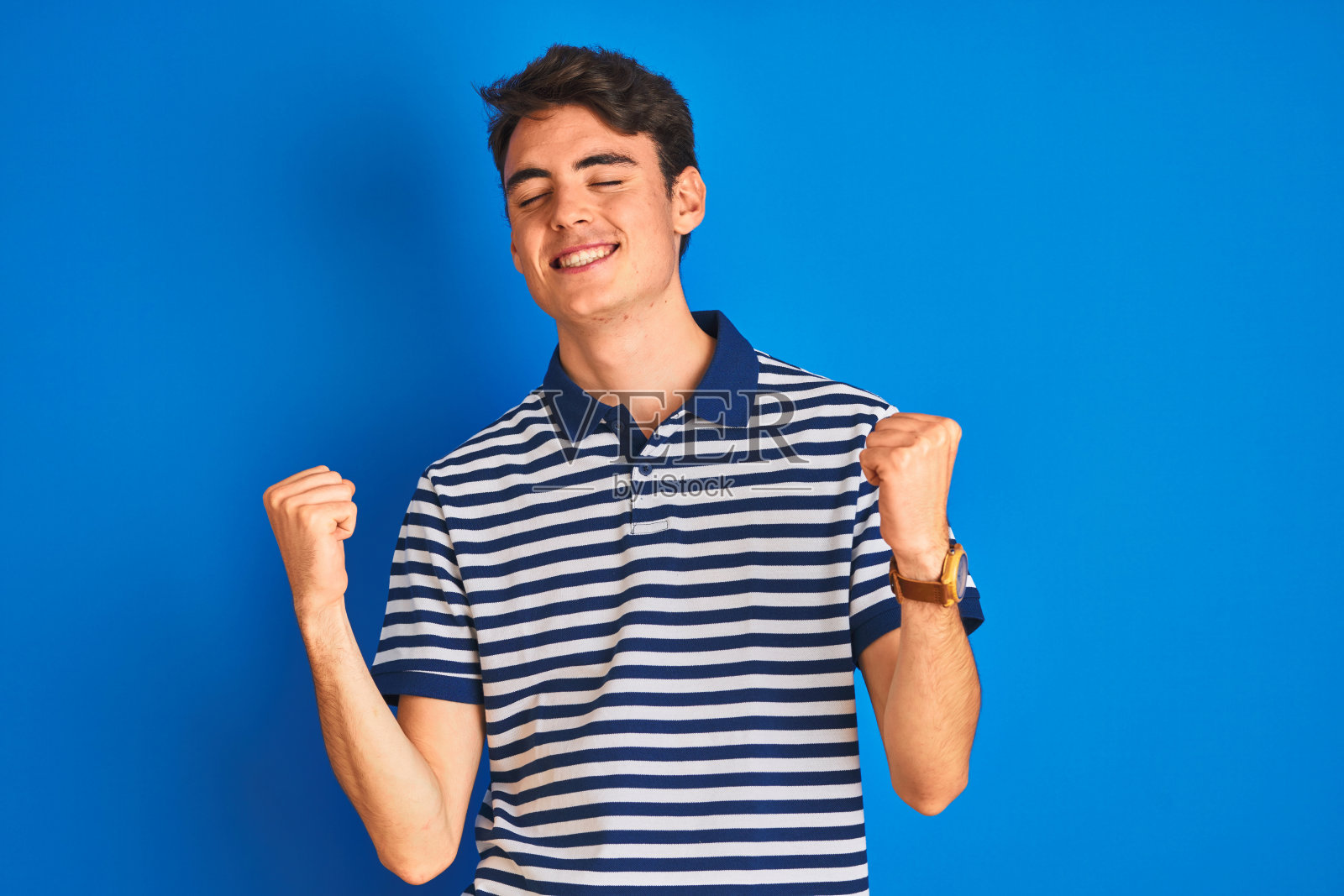 青少年男孩身穿休闲T恤，在蓝色背景前庆祝成功，惊喜和惊讶，举起双臂，闭上眼睛，获胜者概念照片摄影图片