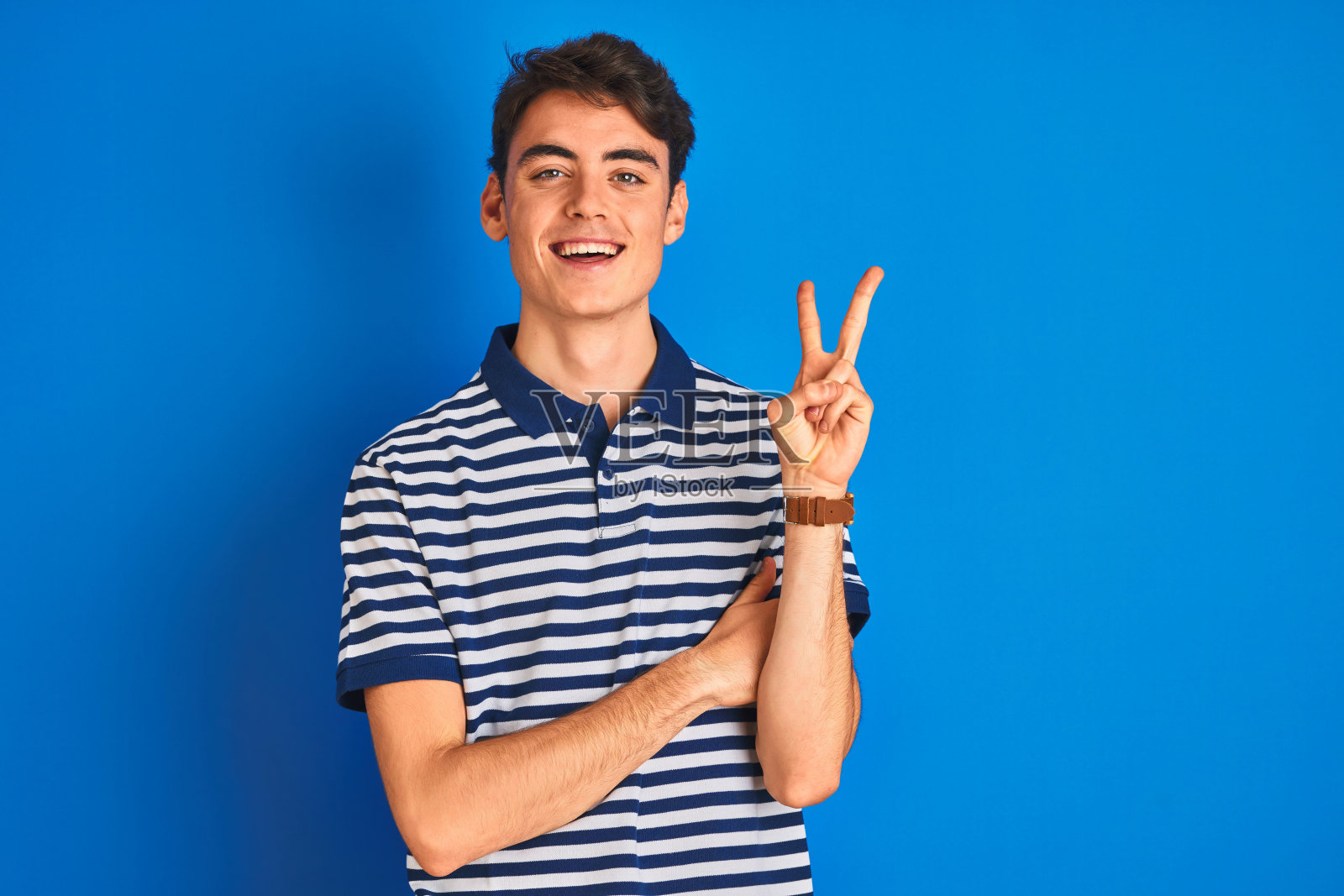 穿着休闲T恤的青少年男孩站在蓝色背景前，面带微笑，向镜头眨眼，用手指做出胜利手势（数字二）照片摄影图片