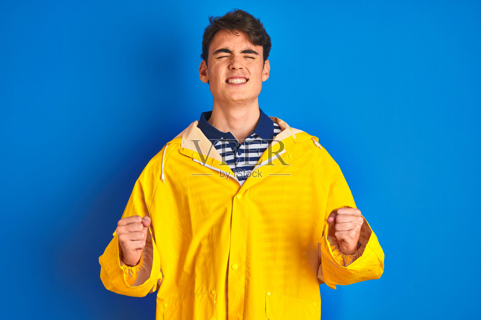 穿着黄色雨衣的少年渔夫，在孤立的背景下，兴奋地举起双臂，闭着眼睛庆祝胜利，面带微笑。 成功者概念。照片摄影图片