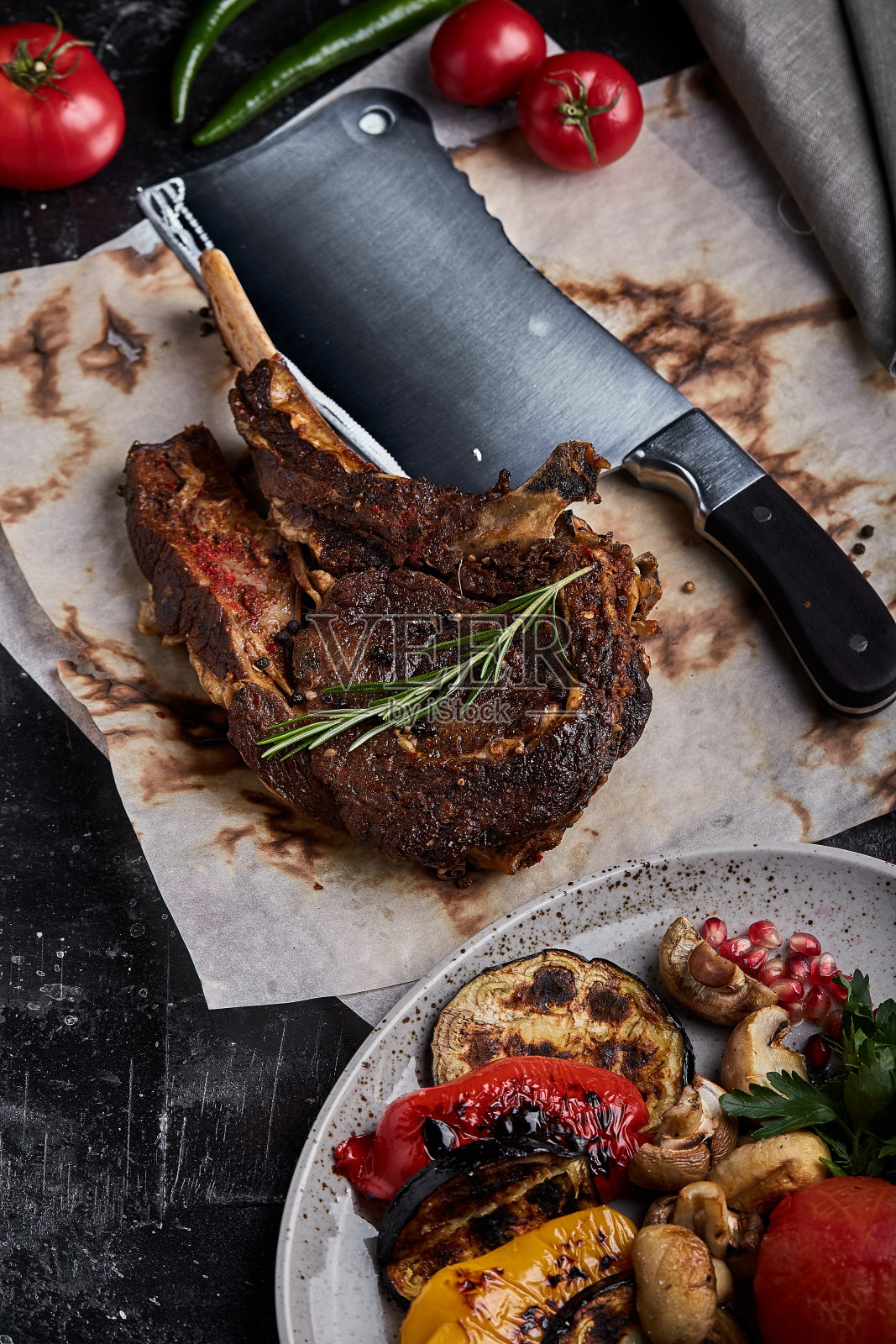 战斧牛排，蔬菜和刀放在桌上。桌上有烤肉、烤蔬菜和新鲜蔬菜照片摄影图片