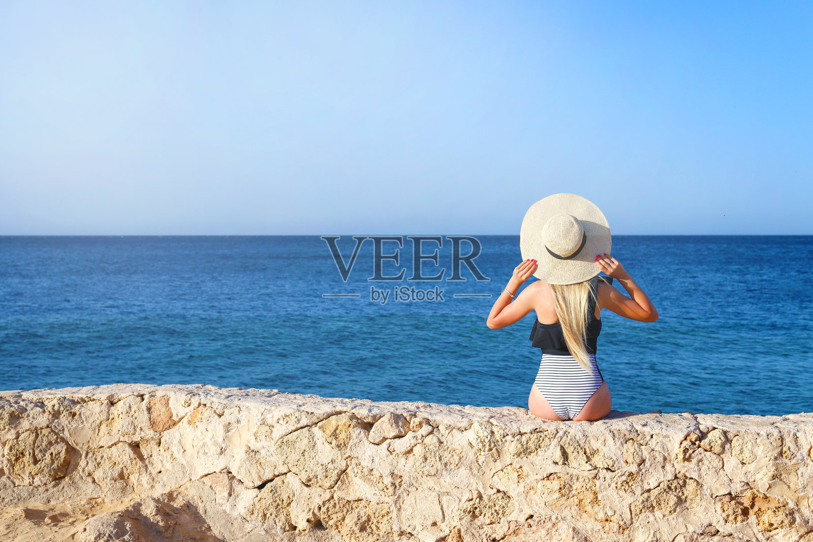 年轻漂亮的女孩穿着泳衣在石头上放松，背景是蓝色的大海和天空照片摄影图片