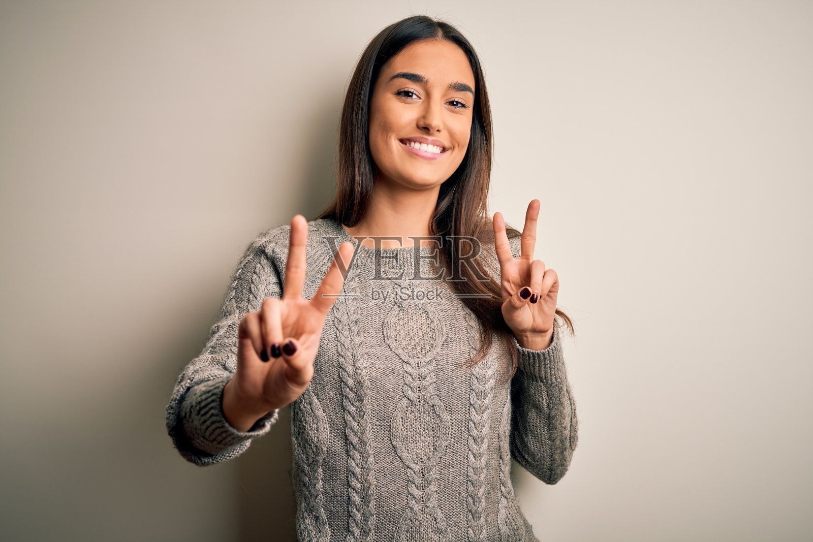 年轻美丽的黑发女人穿着休闲毛衣在孤立的白色背景微笑看着相机显示手指做胜利的手势。二号人物。照片摄影图片