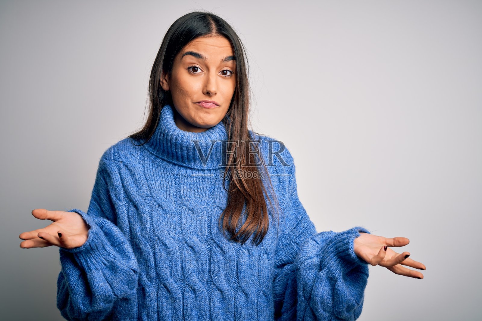 年轻美丽的黑发女人穿着休闲高领毛衣在白色的背景毫无头绪和困惑的表情与胳膊和手举起。怀疑的概念。照片摄影图片