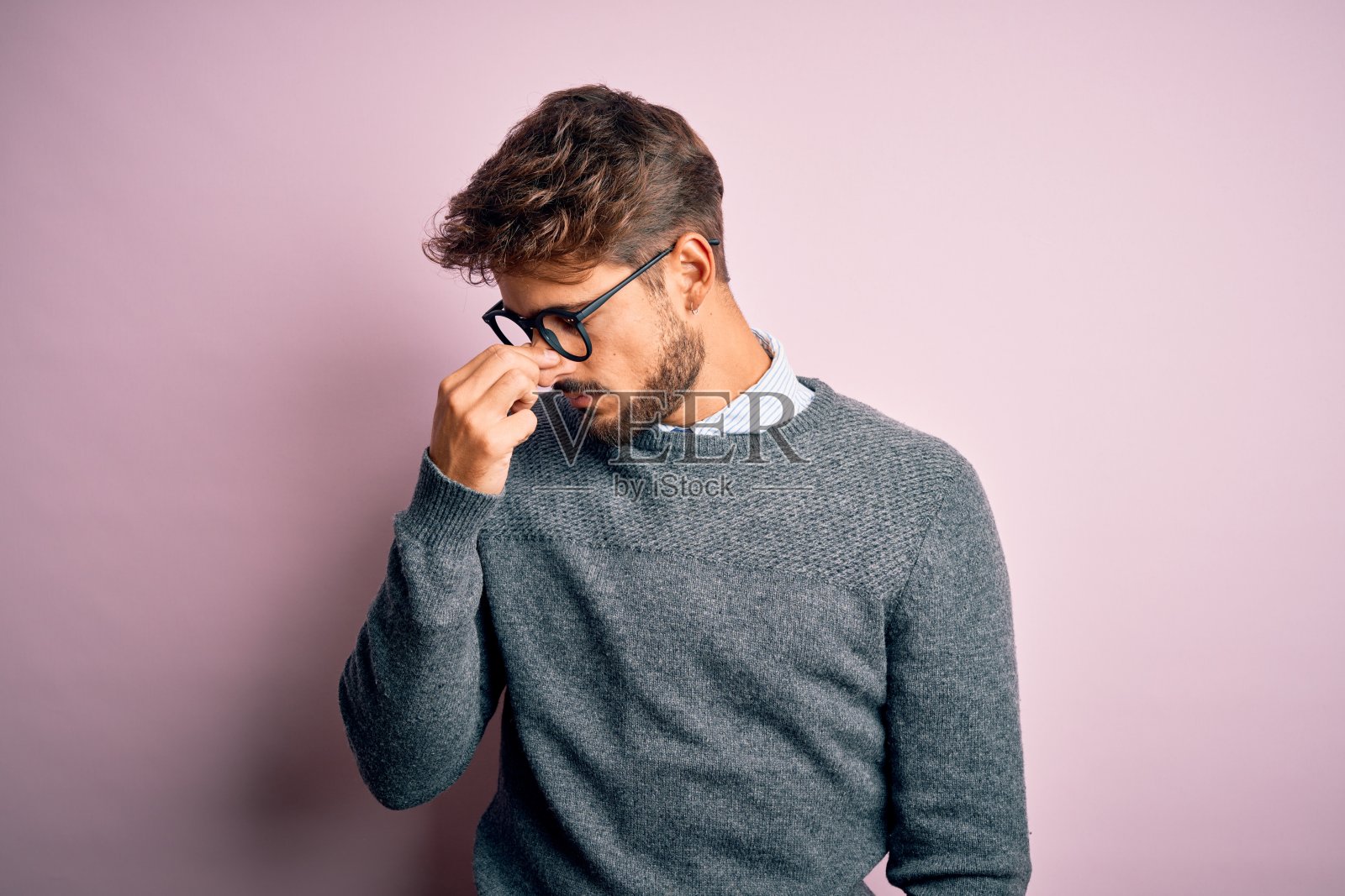 年轻英俊的男子，戴着眼镜和毛衣站在粉红色的背景疲惫揉鼻子和眼睛感觉疲劳和头痛。压力和挫折的概念。照片摄影图片