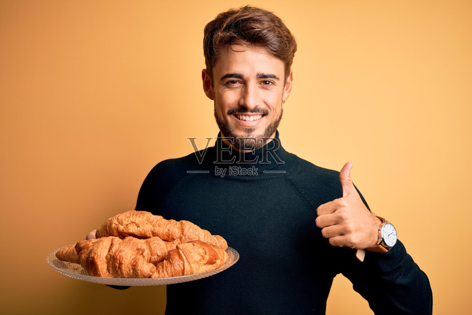 一个留着胡子的年轻人拿着盛有牛角面包的盘子，站在孤立的黄色背景上，开心地笑着做“ok”的手势，用手指竖起大拇指，做“很好”的手势照片摄影图片