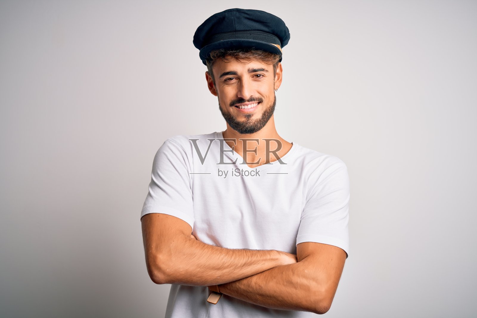 年轻的司机戴着胡子戴着帽子站在孤立的白色背景下，笑脸相迎，双臂交叉看着镜头。积极的人。照片摄影图片