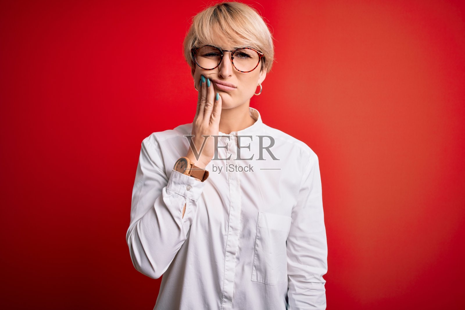 年轻的金发商务女人与短头发戴眼镜在红色的背景触摸嘴与手痛苦的表情，因为牙痛或牙齿疾病的牙齿。牙医照片摄影图片