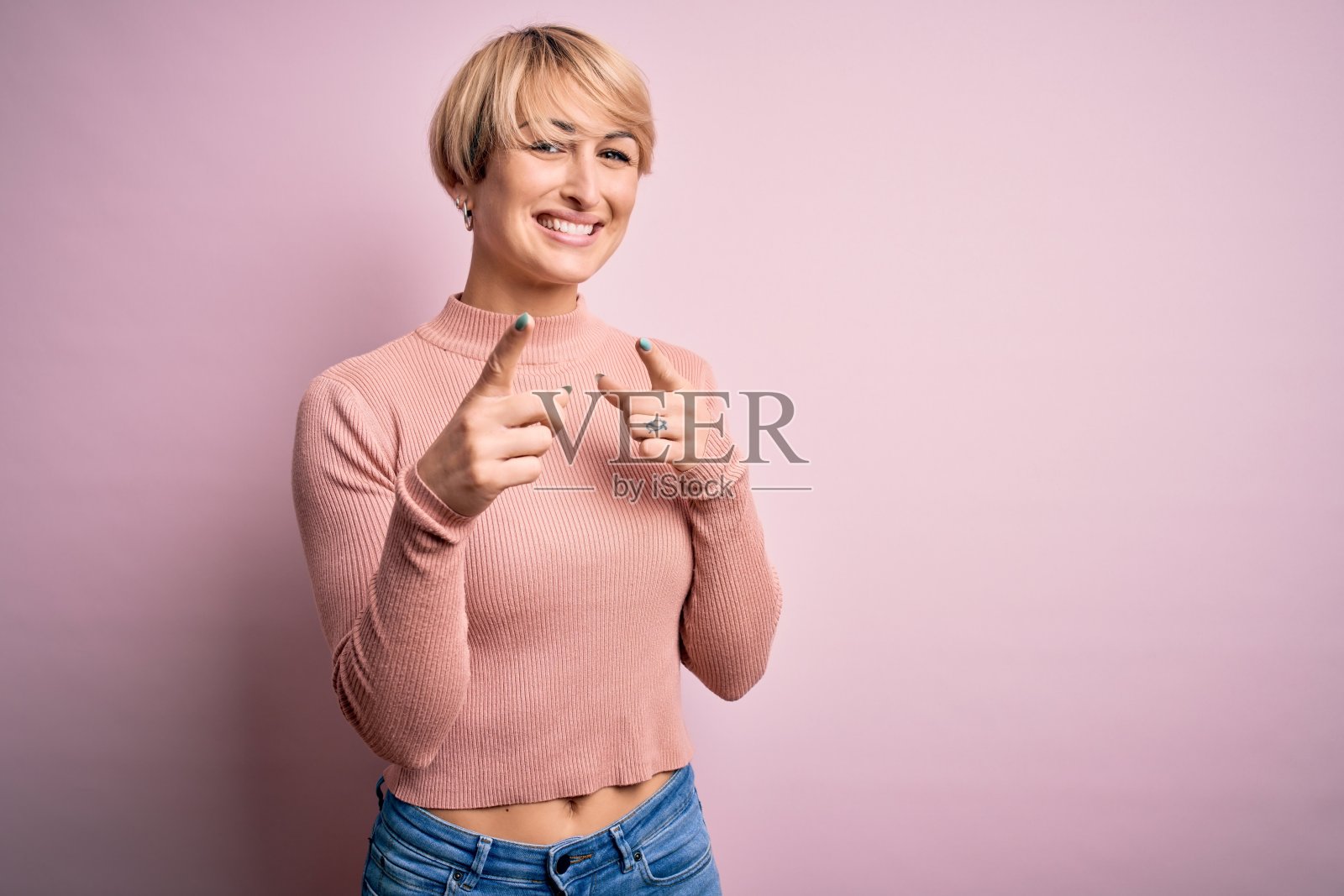 年轻的金发女子与短头发穿着休闲高领毛衣在粉红色的背景指向手指与快乐和有趣的脸相机。良好的能量和共鸣。照片摄影图片