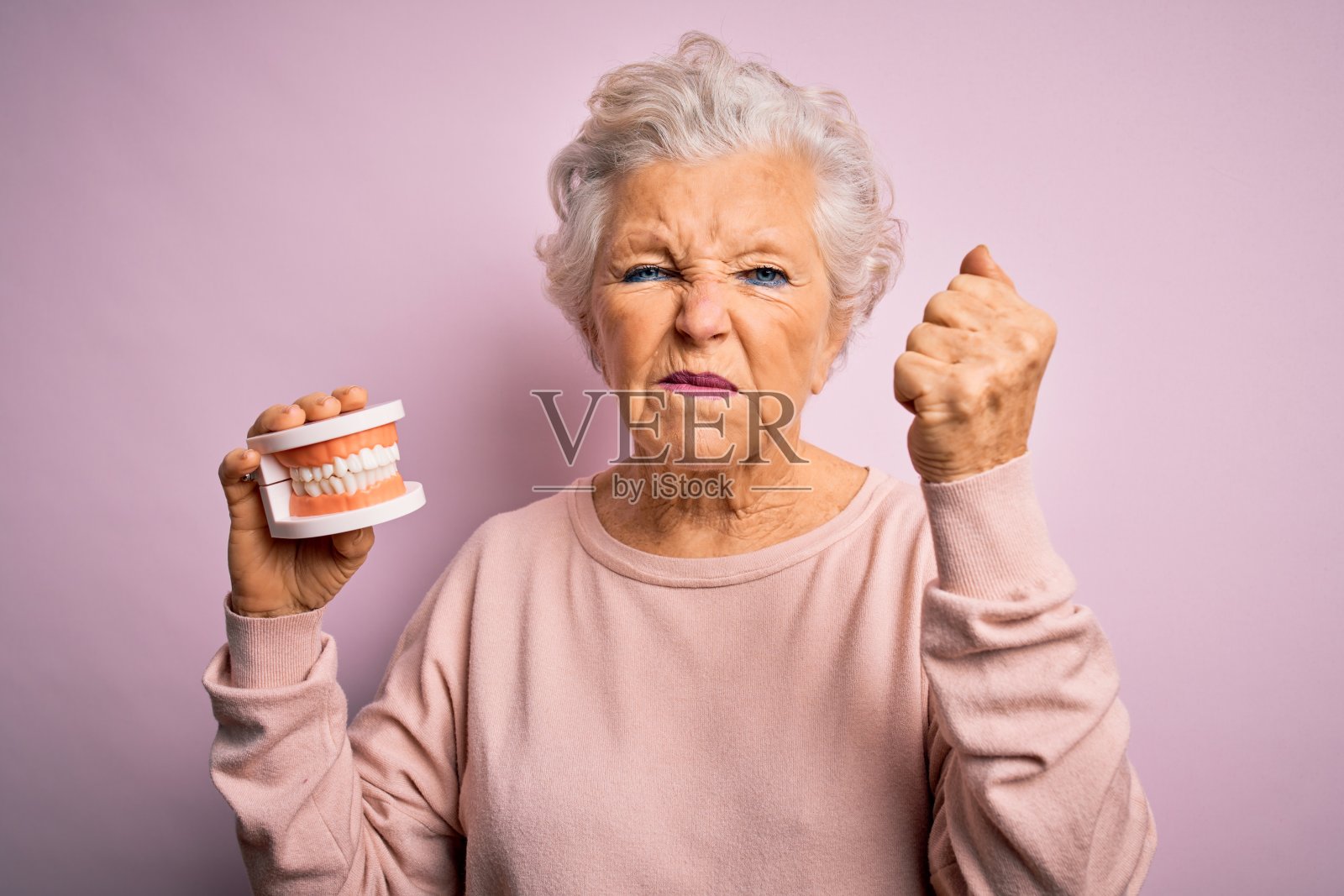 高级美丽的灰白头发的女人拿着塑料假牙在粉红色的背景生气和沮丧的大喊与愤怒，疯狂的大喊与举起的手，愤怒的概念照片摄影图片