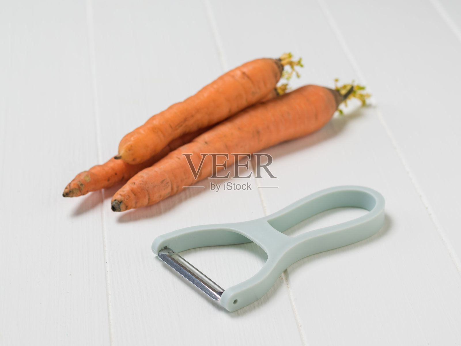 三根胡萝卜和一个削皮器放在一张朴素的白色桌子上。照片摄影图片