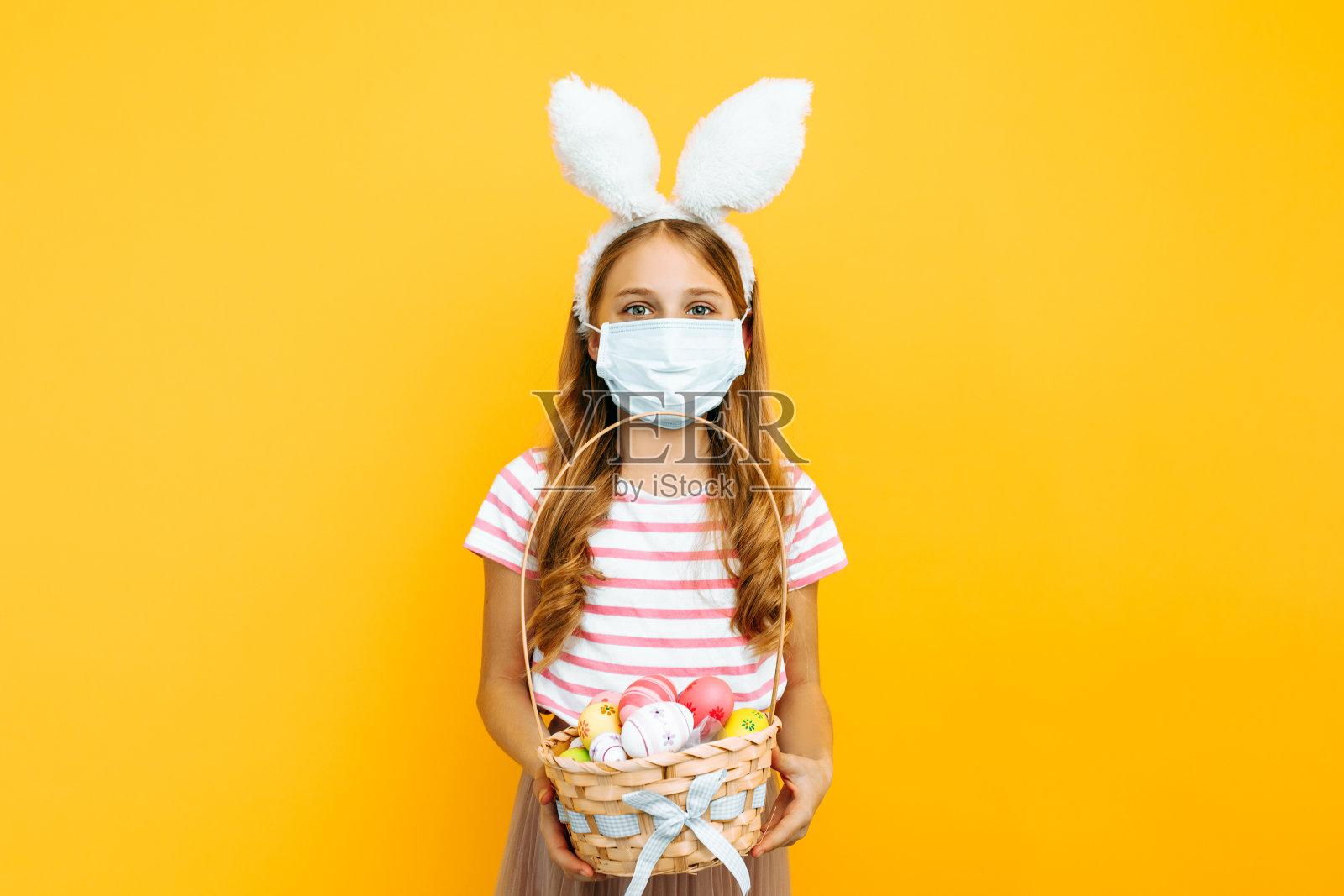 女孩脸上戴着医用冠状病毒口罩，头上戴着兔耳朵，手里拿着一篮子复活节彩蛋照片摄影图片