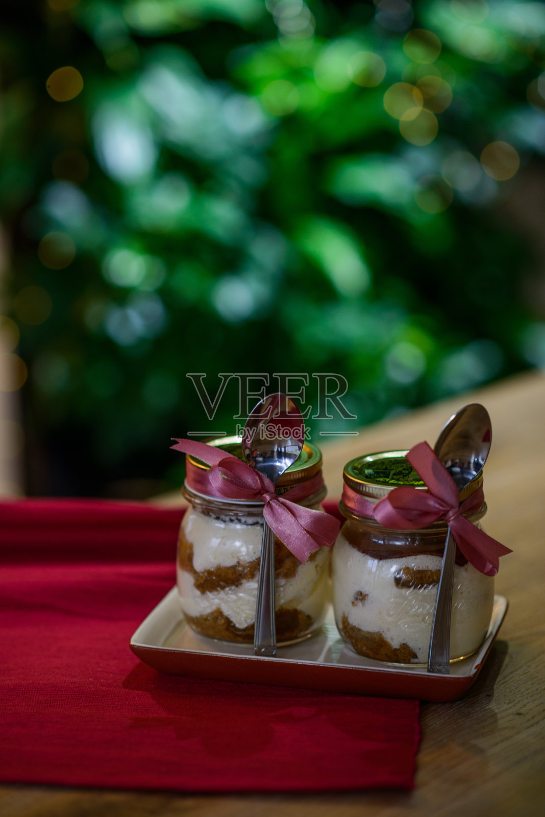 美味的意大利提拉米苏圣诞餐桌用玻璃罐装饰以粉色丝带和银汤匙，在红色餐巾上以灯光和散焦为背景。照片摄影图片