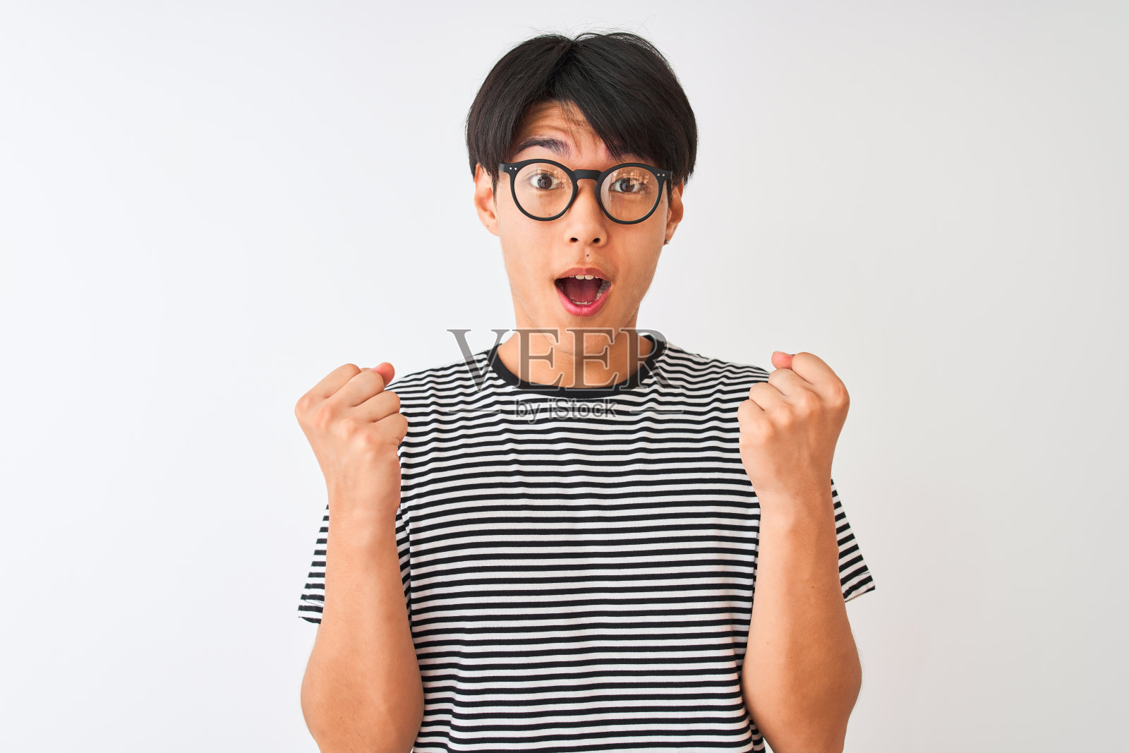 一名中国男子戴着眼镜，身穿海军蓝条纹t恤，站在孤立的白色背景下，举起双臂，睁大眼睛，为成功而惊讶。赢家的概念。照片摄影图片