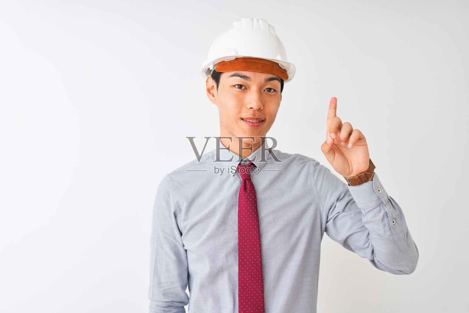 中国建筑师戴着头盔，戴着领带，站在孤立的白色背景下，用第一个手指指着上面，微笑着自信而快乐。照片摄影图片